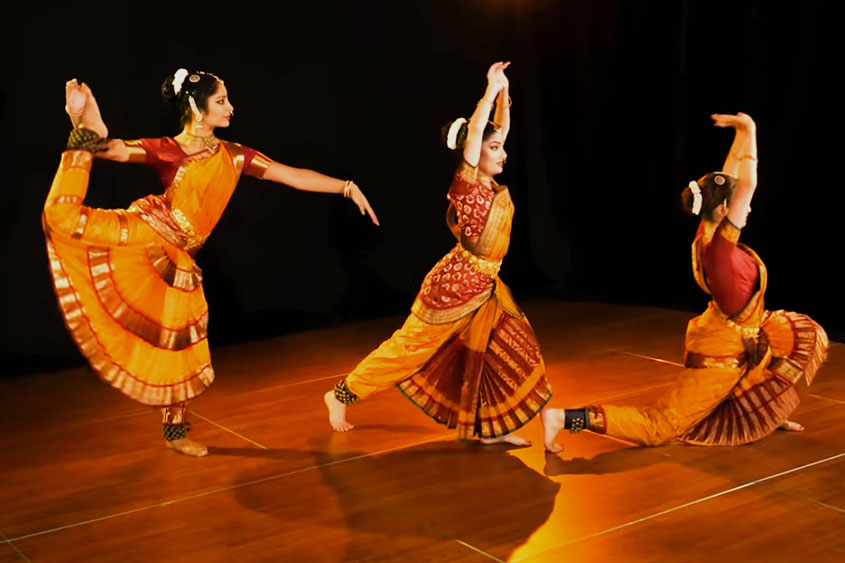 Three Bharatanatyam dancers in golden silk costumes performing synchronized poses on wooden stage