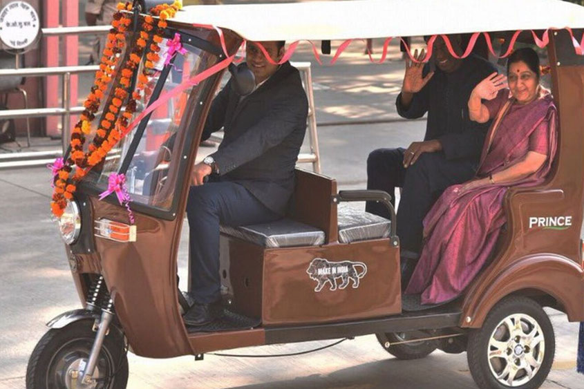 Two officials riding in decorated electric rickshaw adorned with orange and pink flower garlands on paved road.