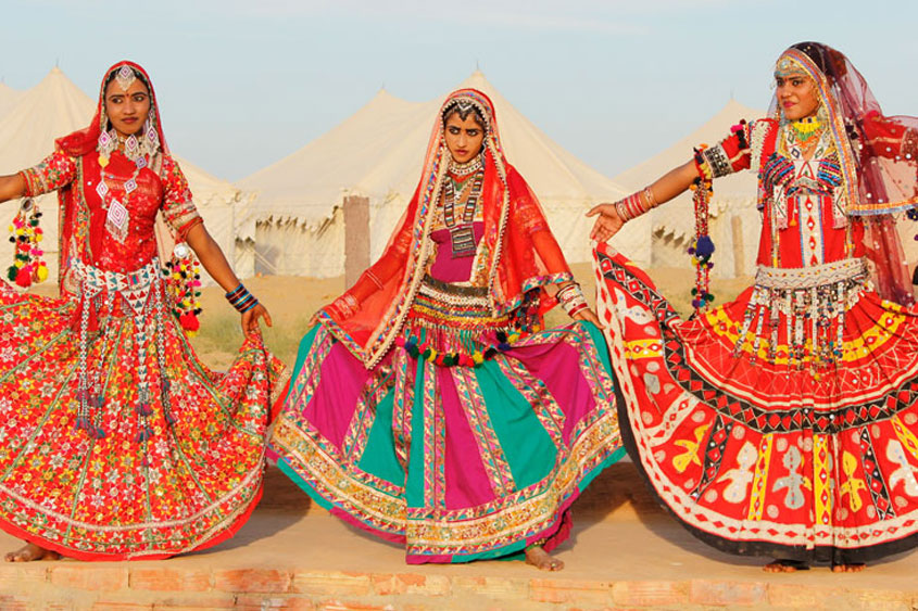 Dancers in folk dance costumes in Rajasthan colors with desert landscape.