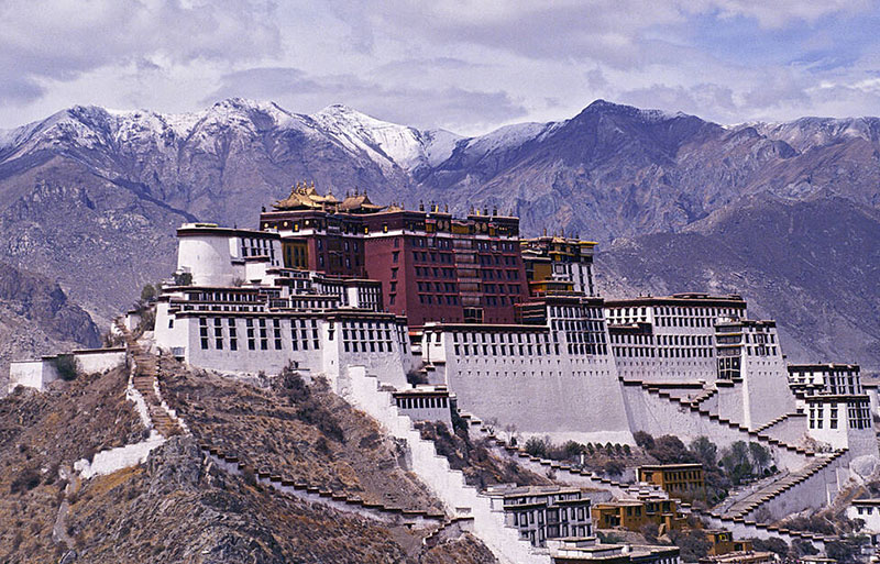 Potala Palace perched on hill with white and red walls against dramatic snow-capped Himalayan mountain range backdrop.