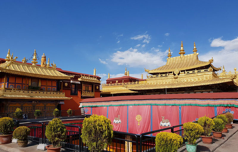Jokhang Temple courtyard with golden roofs, red walls, and ornate Buddhist architecture under clear blue sky.