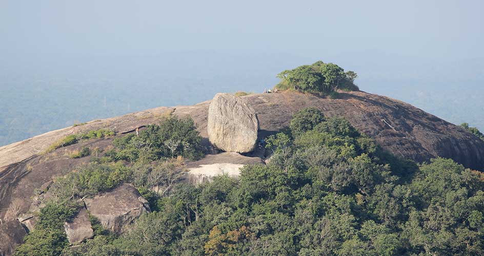 Large balanced boulder atop a forest-covered hill with expansive hazy horizon in the background.