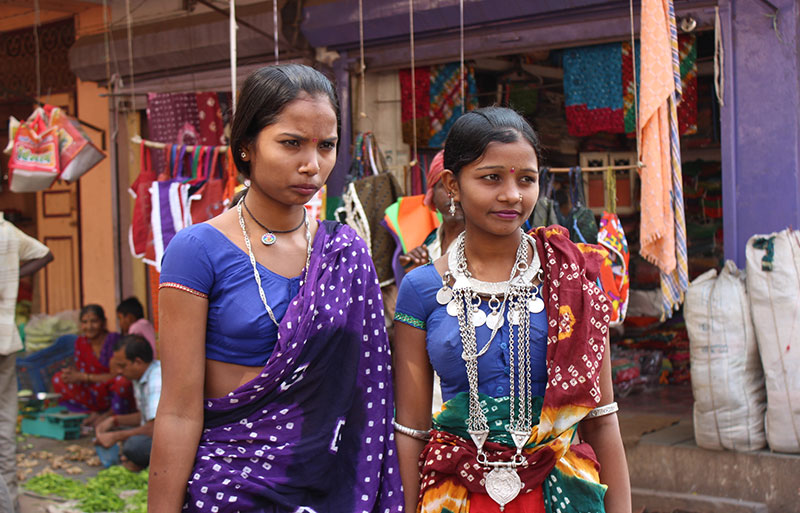 Two women wearing blue tops with traditional silver coin necklaces at market entrance