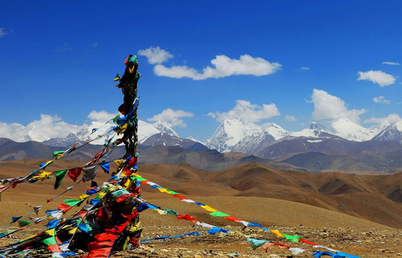 Colorful Tibetan prayer flags on pole overlooking snow-capped mountains and arid brown hills under scattered clouds.