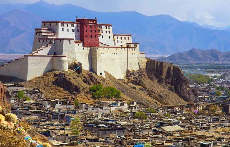 White and red Tibetan fortress perched on rocky hill above town with blue mountains in distant background.