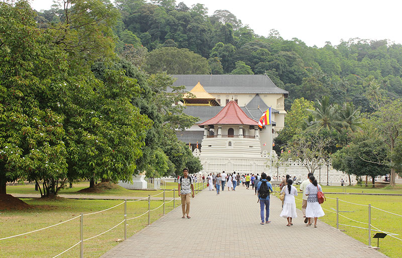 Visitors walking pathway toward Temple of Tooth Relic with red roof amid forested hills