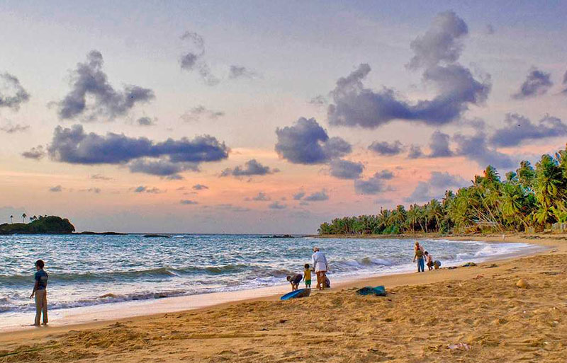 Beach at sunset with people near shoreline, palm-lined coast, and colorful clouds above