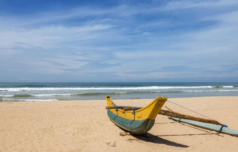 Yellow and blue outrigger fishing boat resting on empty sandy beach with ocean waves