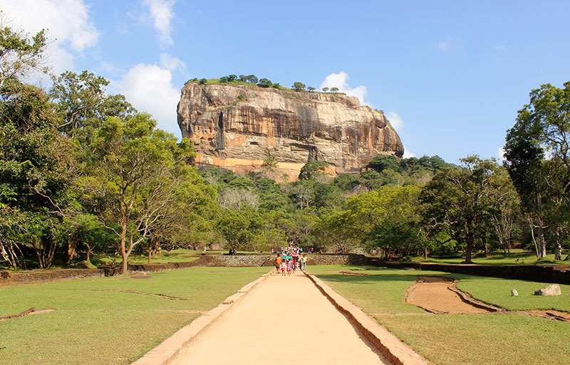 Massive rock fortress rising from gardens with visitors walking pathway toward ancient site