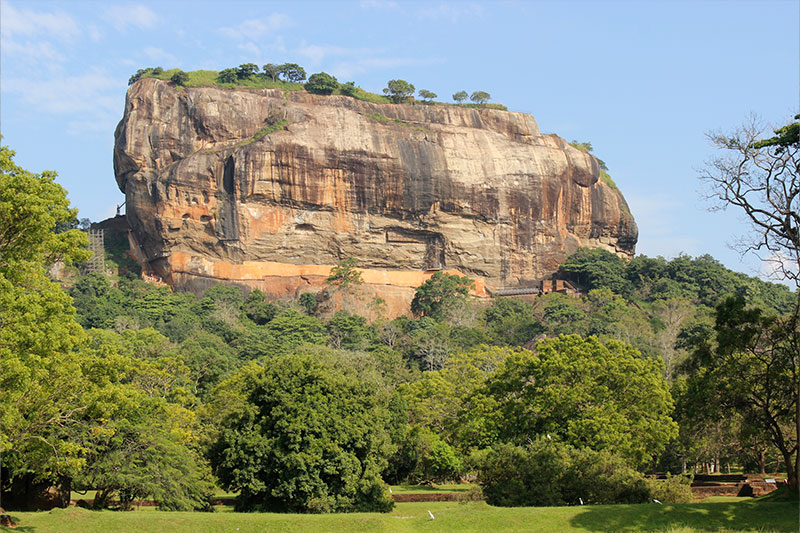 Massive ancient rock fortress rising above lush green forest under clear blue sky in Sri Lanka.