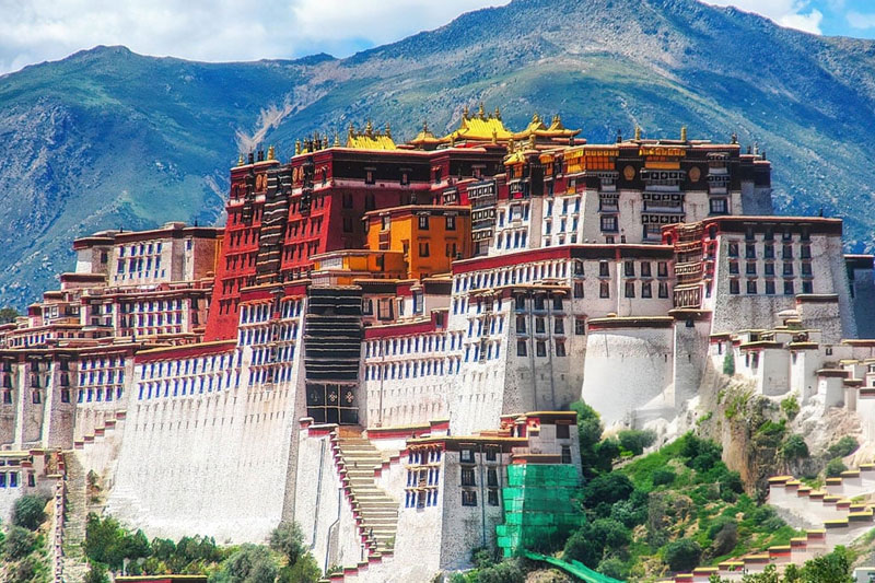 Potala Palace in Lhasa with white, red, and gold buildings rising against blue mountain backdrop under cloudy sky.