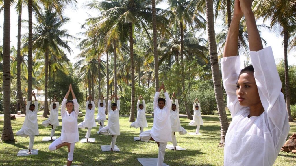 White individuals in a large outdoor yoga practice in the palm tree tropical garden in tree pose.