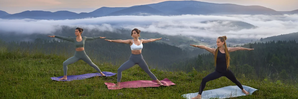 Three women practicing warrior two yoga pose on mountain meadow with misty forest valley below.